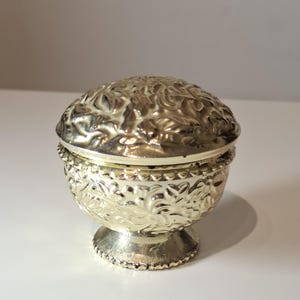 Pode incluir: Pequena caixa de bugigangas dourada com tampa. A caixa apresenta um padrão floral detalhado e assenta numa pequena base de pedestal. A tampa é arredondada e também apresenta o mesmo design floral.
