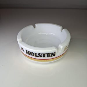 Puede incluir: Cenicero de cerámica blanca con forma redondeada y cuatro muescas para apoyar cigarrillos. El cenicero presenta la palabra "HOLSTEN" en letras negras, con un diseño de rayas doradas y rojas. Un accesorio clásico para fumadores.
