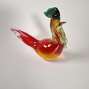 Könnte beinhalten: Eine Glasfigur eines Hahns mit einem rot-gelben Körper, einem langen roten Schwanz und einem Hals mit wirbelnden grünen, gelben und klaren Glasakzenten. Der Kopf ist mit einem dunkelgrünen Kamm versehen und der Schnabel zeigt nach oben.
