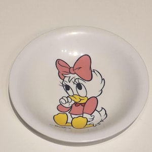 May include: White ceramic bowl with a cartoon image of Daisy Duck. The bowl features Daisy with a pink bow, dress, and yellow feet. The round bowl has a simple design, suitable for serving food, with a diameter of approximately 20 cm.