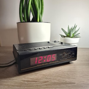 Puede incluir: Reloj despertador digital negro que muestra las 12:05 en rojo. El reloj tiene un gráfico de mapa del mundo y el texto "AM/FM Radio Clock". Una planta verde en una maceta blanca y una pequeña suculenta están en el fondo.
