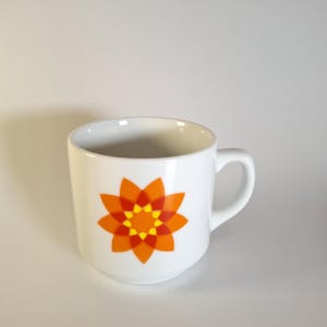 Può includere: Tazza in ceramica bianca con un vivace motivo floreale arancione e giallo. La tazza ha un manico curvo e un design semplice e pulito. Il motivo floreale è simmetrico.