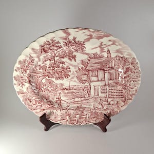 Può includere: Piatto ovale in ceramica rossa e bianca con bordo smerlato. Il piatto presenta una scena pastorale con una casa, alberi e figure. È esposto su un supporto in legno marrone.