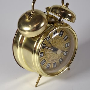 Könnte beinhalten: Ein goldfarbener Metall-Doppelglockenwecker mit rundem Zifferblatt. Die Uhr hat schwarze römische Ziffern auf einem goldenen, gemusterten Hintergrund, einen Tragegriff und zwei Glocken oben. Das Wort "CUPPER" ist schwarz auf dem Zifferblatt gedruckt.