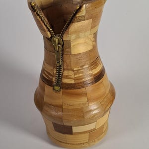 Pode incluir: Um vaso construído com pequenos blocos de madeira de vários tons dispostos num padrão de mosaico. A secção superior apresenta um fecho de correr metálico cor de latão integrado na madeira, descendo do rebordo para o centro.