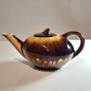 Könnte beinhalten: Eine braune und beige Keramik-Teekanne mit Deckel und Henkel. Die Teekanne hat einen abgerundeten Körper mit Ausguss und gebogenem Henkel. Der Farbverlauf geht von Dunkelbraun zu Beige über.