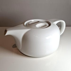 Può includere: Una teiera in ceramica bianca con corpo arrotondato, manico curvo e beccuccio. Il coperchio ha un'indentazione circolare. La teiera è su una superficie bianca.