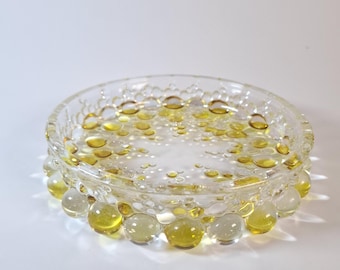 Può includere: Un piatto circolare in vetro con una superficie strutturata caratterizzata da numerose bolle sferiche in rilievo. Il design incorpora un mix di sfere in vetro trasparente e sfere color giallo traslucido, creando un motivo a pois.