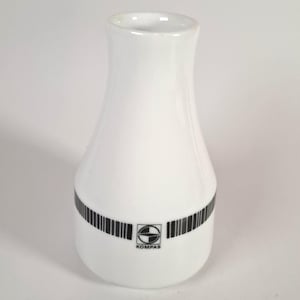 May include: White ceramic vase with a flared top and a rounded base. A black barcode-like design encircles the base, with the word "KOMPAS" and a logo in the center. The vase has a smooth, glossy finish.