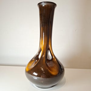 May include: A tall, brown vase with an unusual, organic form. The vase features a rounded base that narrows to a slim neck and a flared top. The colour shifts from a dark brown to a lighter amber shade, producing a striking visual effect.