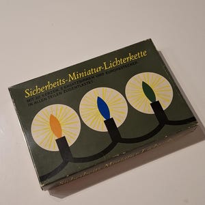 May include: A vintage box with the text "Sicherheits-Miniatur-Lichterkette" in German. The box features a design of three stylized candles with orange, blue, and green flames. The box is dark green with a white background.