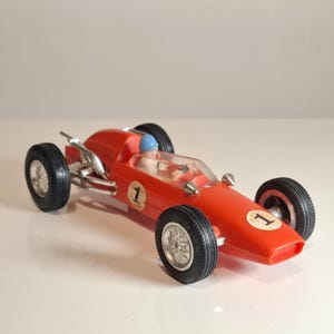 Pode incluir: Um carro de corrida de brinquedo vermelho vintage com pneus pretos e aros prateados. O carro tem o número 1 na lateral e na frente. Um pequeno piloto com capacete azul é visível dentro. Um carro de brinquedo clássico.