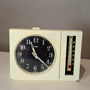 Può includere: Sveglia vintage color crema con quadrante nero, numeri bianchi e lancette verdi. L'orologio include un termometro con indicatore rosso. Il quadrante dell'orologio indica "Insa" e "Yugoslavia".