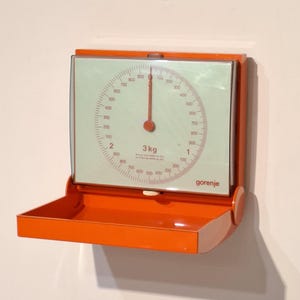 Può includere: Una bilancia da cucina vintage arancione con un quadrante circolare e un piccolo vassoio. Il quadrante ha segni rossi e un'ago rosso, con la scritta "3 kg" e il marchio "gorenje". La bilancia è montata a parete.