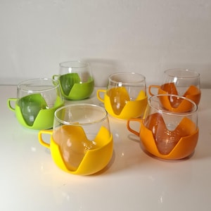 Könnte beinhalten: Sechs klare Teetassen aus Glas mit farbigen Silikonhaltern in Grün, Gelb und Orange. Jede Tasse hat einen kleinen Henkel und einen gebogenen Silikonboden. Die Tassen sind auf einer weißen Oberfläche angeordnet.