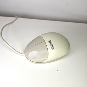Op de afbeelding: Een crèmekleurige computermuis met een lichtgrijze accentkleur en een witte kabel. Het woord "SIGMA" staat in blauw op de zijkant. De muis heeft een afgeronde vorm met een ronde knop.