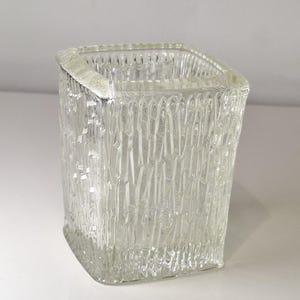 Può includere: Vaso quadrato in vetro trasparente con un motivo verticale strutturato. Il vaso ha una forma quadrata leggermente arrotondata e un bordo spesso. Il vetro sembra pesante e resistente, adatto per contenere fiori o altri oggetti decorativi.