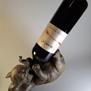 Op de afbeelding: Een bruine olifantvormige wijnfleshouder ondersteunt een donkere wijnfles. De fles heeft een wit etiket met rode accenten en tekst. De olifant ligt op zijn rug en houdt de fles vast met zijn slurf en slagtanden. Het is een decoratief item.