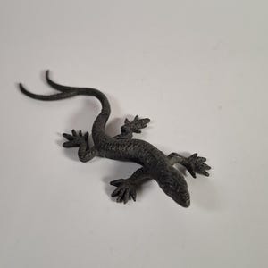 Op de afbeelding: Een donkergrijs, getextureerd metalen hagedisbeeldje met een lange, slanke staart en vier gespreide poten. Het oppervlak van de hagedis is voorzien van ingewikkelde, verhoogde schubachtige patronen over het hele lichaam, de kop en de ledematen.