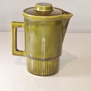 以下が含まれることがあります： 蓋とハンドルが付いたオリーブグリーンのセラミックコーヒーポット。ポットは、リブ付きのベースと注ぎ口を備えた円筒形のボディをしています。蓋の上部には小さなノブが付いています。