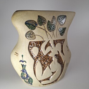 Puede incluir: Un jarrón de cerámica beige con una forma asimétrica inusual. El jarrón presenta diseños marrones, texturizados y tallados, incluyendo hojas estilizadas y un elemento decorativo en el lateral. Las hojas tienen acentos verdes y azules.