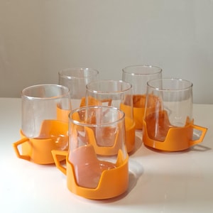 Pode incluir: Conjunto de sete canecas de vidro transparente com suportes de plástico laranja. As canecas têm formato cilíndrico e são projetadas para se encaixarem perfeitamente nos suportes laranja, que possuem uma alça. As canecas estão dispostas sobre uma superfície branca.