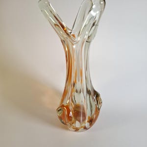 May include: A clear glass vase with an organic, flowing shape. The vase has a flared top with three upward-reaching sections. Amber and orange hues are swirled throughout the glass, creating a decorative effect. The vase is a decorative home decor item.