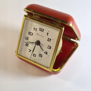 Op de afbeelding: Een vintage reiswekker met een rode kunstleren etui en goudkleurige randen. De wijzerplaat is wit met zwarte cijfers en wijzers, en het woord "Blessing" staat op de wijzerplaat.