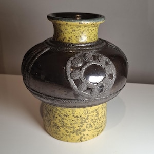 May include: A ceramic vase with a bulbous, dark brown center and a speckled yellow base and neck. The center features a raised, circular design with a silver-colored center. The rim is dark green. The vase has a unique, textured appearance.