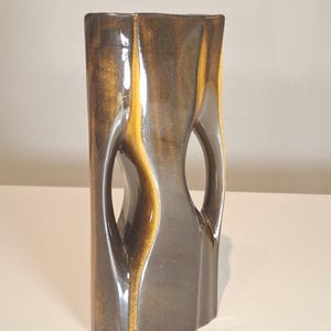 Op de afbeelding: Een hoge, rechthoekige keramische vaas met een uniek ontwerp. De vaas heeft een donkerbruine en goudkleurige glazuur, met twee ovale uitsparingen aan de zijkanten. De vaas heeft een glanzende afwerking en is geschikt voor woondecoratie.