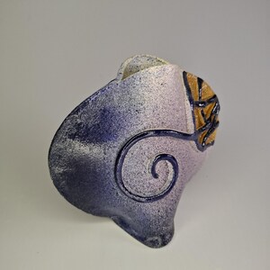 May include: A ceramic vase with a unique design. The vase has a speckled blue and white exterior with a dark blue spiral accent. A section of the vase has a textured, golden-brown leaf design. The vase has an organic, asymmetrical shape.