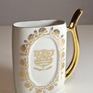 May include: White ceramic mug with a gold handle and gold floral detailing. The mug has an oval design with the words "KARLOVY VARY" and a gold emblem. The handle is a curved, solid gold colour.