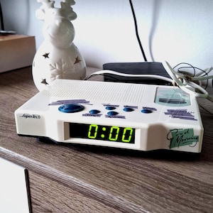 Pode incluir: Um despertador digital branco Supertech com um visor LED verde mostrando 0:00. Possui botões azuis e as palavras "Good Morning" na lateral. Uma figura de veado de cerâmica branca está no fundo.