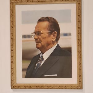 Op de afbeelding: Een ingelijst portret van een ouder persoon in profiel, gekleed in een donker pak, een wit overhemd met kraag en een gestreepte das. De persoon heeft kort bruin haar en draagt een bril met een licht montuur. De lijst is van licht, gestructureerd hout.