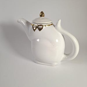 Pode incluir: Um bule de cerâmica branca em forma de gato com uma alça curva e um bico. O bule possui uma tampa removível com um remate dourado e um detalhe de laço pintado em ouro ao redor da área do pescoço, proporcionando um destaque decorativo.