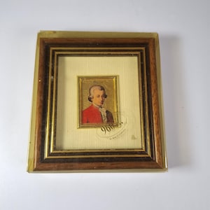 Op de afbeelding: Een rechthoekig ingelijst portret van Wolfgang Amadeus Mozart met een inleg van goudfolie. Het portret is geplaatst tegen een crèmekleurige, gestructureerde achtergrond in een donkerhouten lijst met gouden accenten en een doorzichtige plastic hoes.