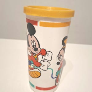 Puede incluir: Vaso de plástico blanco con tapa amarilla, con los personajes de dibujos animados Mickey y Minnie Mouse. El vaso tiene rayas rojas, naranjas y verdes. Un objeto de colección divertido y colorido.