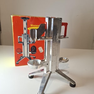 以下が含まれることがあります： 赤色のハンドルが付いた、シルバーメタルのヴィンテージWestmark柑橘類ジューサー。ジューサーは三本足のベースとボウル型のレセプタクルを備えています。ジューサーのイラストが描かれたオリジナルの箱が背景に見えます。