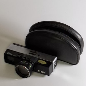 Op de afbeelding: Een zwarte en zilveren PORST camera met een lens en een zwarte leren etui. De camera heeft een zilveren bovenkant en een zwarte body met een lens en een groene sticker met "PASSED". De camerahoes is ovaal van vorm.