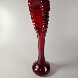 May include: A tall, deep red glass vase featuring a flared, fan-shaped top with a ribbed, shell-like texture. The vase has a slender, smooth neck that transitions into a rounded, bulbous base. A small, circular sticker is visible on the lower body.