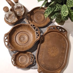 May include: A selection of brown wooden serving dishes and a condiment set. The dishes have carved geometric and floral patterns. The condiment set includes two glass bottles with wooden stoppers in a wooden carrier. A decorative plant is in the background.