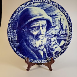 Può includere: Un piatto decorativo blu e bianco con un ritratto dettagliato di un uomo anziano con la barba, che indossa un cappello e fuma la pipa. Lo sfondo raffigura una scena costiera con barche a vela. Il piatto è presentato su un supporto di legno marrone.
