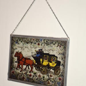 Può includere: Un pannello rettangolare in vetro colorato raffigurante una carrozza trainata da cavalli. La carrozza è gialla e nera, trainata da due cavalli marroni. Il pannello è incorniciato con un metallo argentato e ha un bordo floreale decorativo.