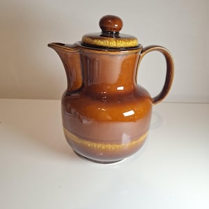 Puede incluir: Una cafetera de cerámica marrón con tapa y asa. La jarra tiene un acabado brillante con una parte superior marrón oscuro y una banda marrón claro alrededor del centro. La tapa tiene un pequeño pomo redondo.