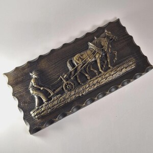 Può includere: Targa da parete rettangolare in legno marrone scuro con una scultura a rilievo raffigurante un contadino che ara un campo con due cavalli. I cavalli e il contadino sono di colore oro e i bordi della targa sono smerlati.