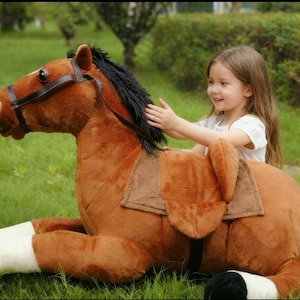 May include: A large, plush brown horse toy with a black mane and tail, and white accents on the legs. The toy has a saddle and bridle. A child is sitting on the toy in a grassy area.