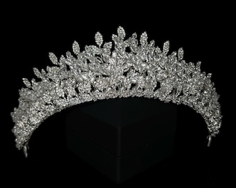 May include: A silver-toned tiara with a leaf-like design, set with numerous clear crystals. The tiara is arched and sits atop a black surface, with a black background. The crystals sparkle, reflecting light.