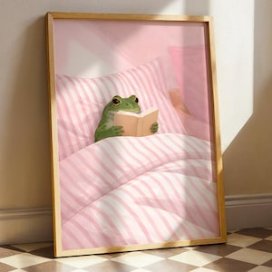 Peut inclure: Une affiche encadrée représente une grenouille verte lisant un livre au lit. La grenouille est sous une literie et des oreillers à rayures roses et blanches. L'œuvre a un cadre en bois clair et est appuyée contre un mur.