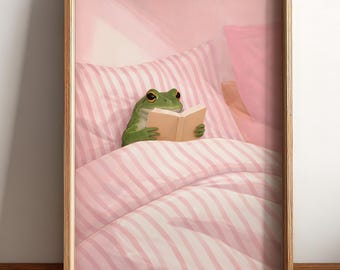 May include: A framed wall art print featuring a green frog lying in a bed with pink and white striped bedding. The frog is holding an open book with both hands, resting against a matching striped pillow in a soft, light-filled bedroom setting.