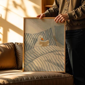 May include: A framed art print featuring a whimsical illustration of a white duck reading a book in bed. The artwork has a soft color palette with blue and beige tones, and a wooden frame.